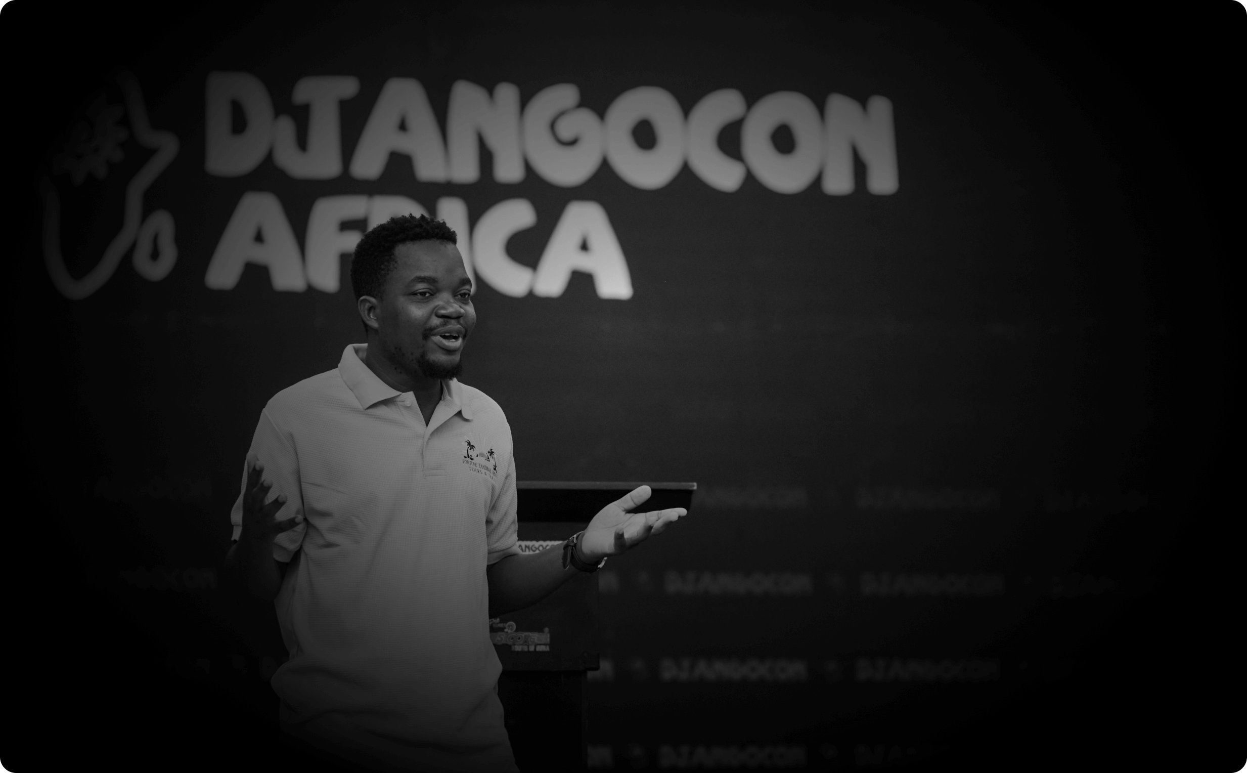 A photo of Hope Adoli on a stage at Djangocon Africa, 
          giving a talk. The photo is in black and white with DjangoCon 
          Africa logo on a 10 x 20 banner blurred behind him